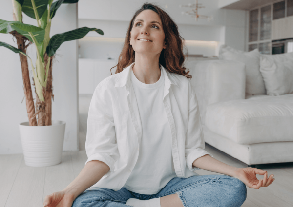 A woman sitting cross-legged on the floor in a bright living room, smiling and practicing meditation near a large indoor plant.